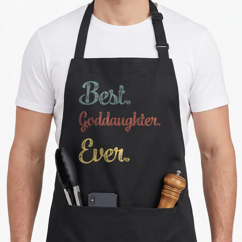 Funny goddaughter apron - Best. Goddaughter. Ever. text in blue-grey, coral, and gold stacked typography — shown on a black cotton canvas apron against a white background