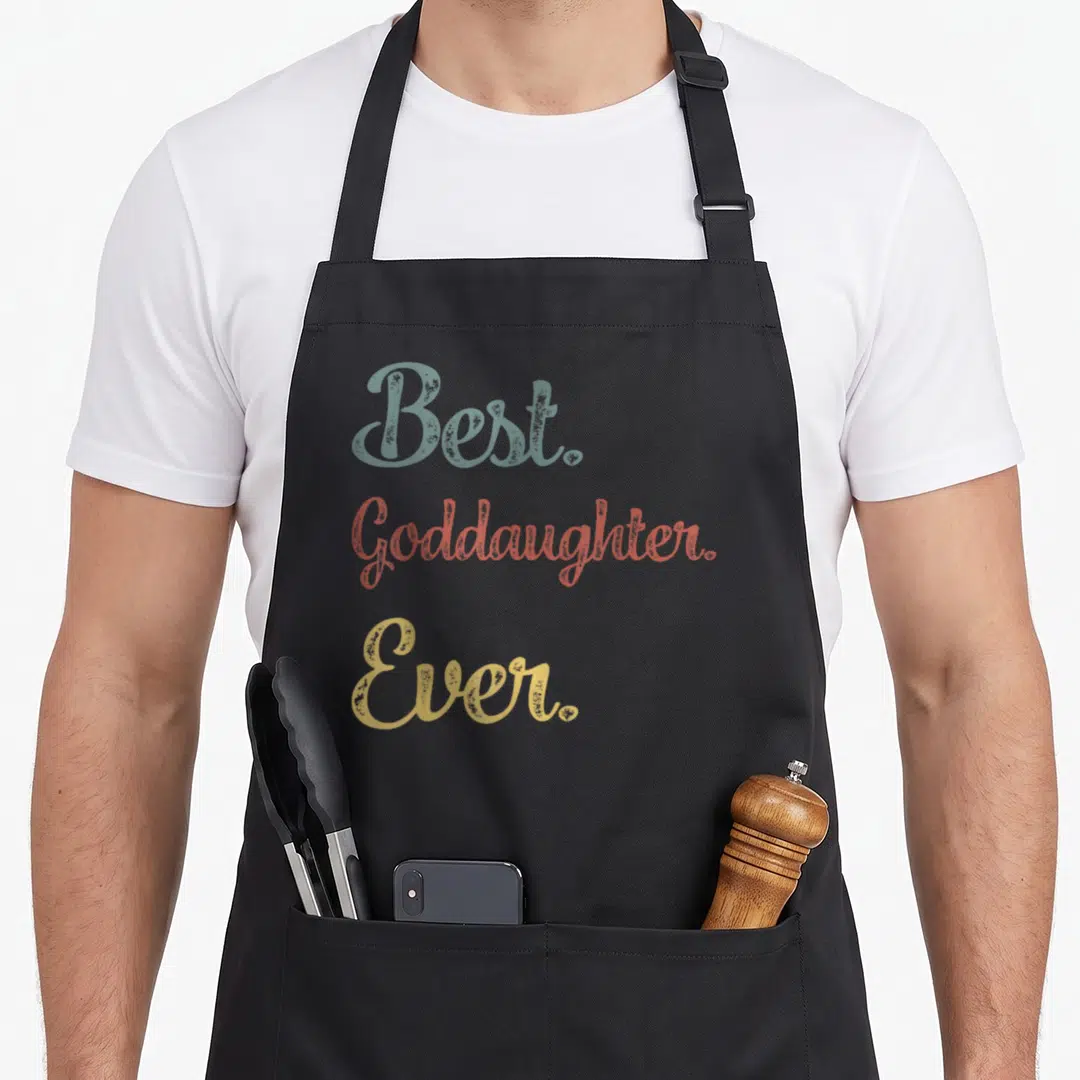 Funny goddaughter apron - Best. Goddaughter. Ever. text in blue-grey, coral, and gold stacked typography — shown on a black cotton canvas apron against a white background