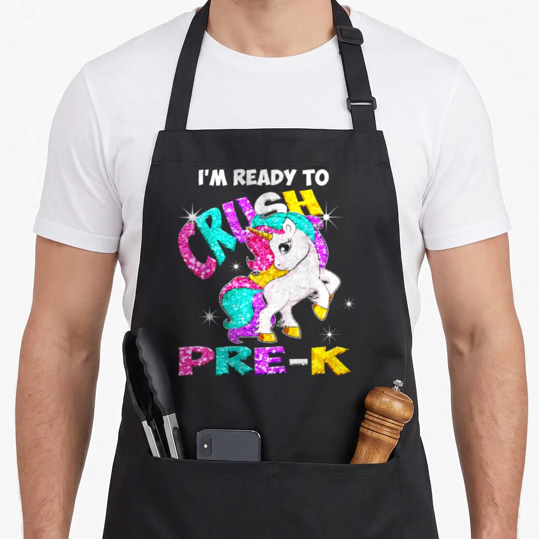 School ready unicorn apron - Cartoon unicorn with rainbow mane and I'M READY TO CRUSH PRE-K text with white stars — shown on a black cotton canvas apron against a white background