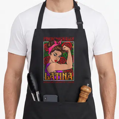 Latin hispanic aprons - Cartoon woman flexing arm inside white frame with PHENOMENALLY LATINA text and rose background — shown on a black cotton canvas apron against a white background