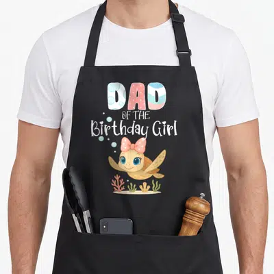 Sea turtle birthday apron - DAD OF THE Birthday Girl text above a cartoon turtle with a pink bow swimming over coral — shown on a black cotton canvas apron against a white background