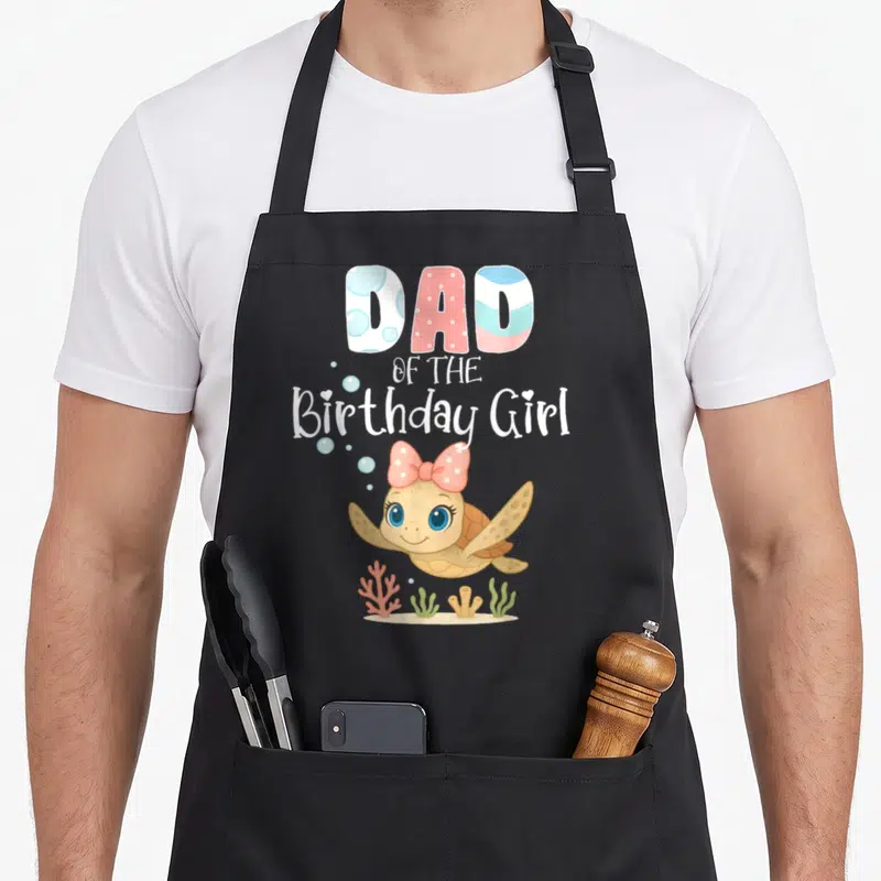 Sea turtle birthday apron - DAD OF THE Birthday Girl text above a cartoon turtle with a pink bow swimming over coral — shown on a black cotton canvas apron against a white background
