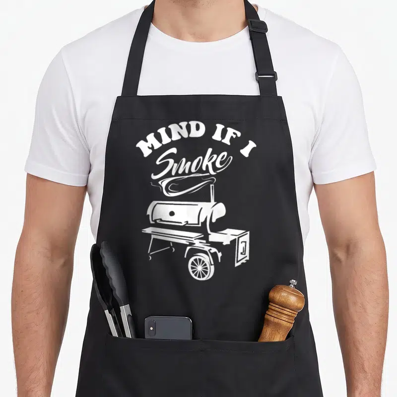 Mind smoker apron - White text MIND IF I Smoke arched over a smoker grill with curling smoke lines — shown on a black cotton canvas apron against a white background