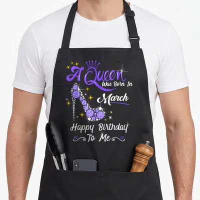 March birthday baking queen apron - Purple high heel shoe with A Queen Was Born In March text and crown graphic — shown on a black cotton canvas apron against a white background