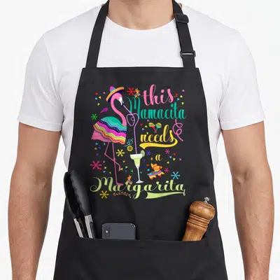 Mamacita pink flamingo apron - Pink flamingo in sombrero sipping margarita with colorful text and confetti — shown on a black cotton canvas apron against a white background