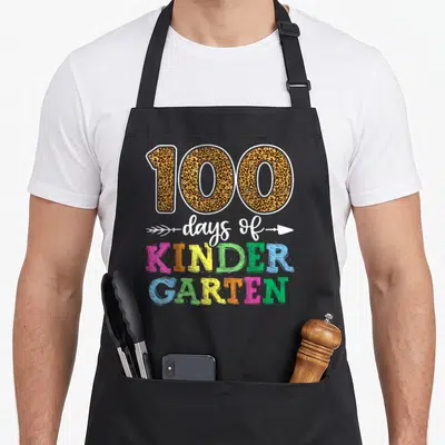 Cute school kindergarten teacher apron - Leopard print 100 with white arrow script and stacked pink yellow green blue — shown on a black cotton canvas apron against a white background
