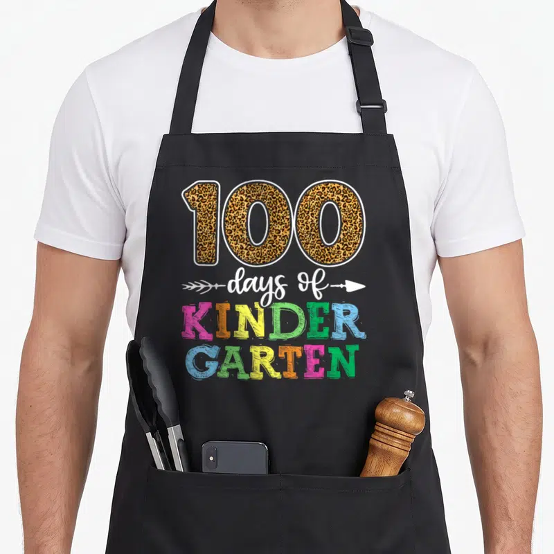 Cute school kindergarten teacher apron - Leopard print 100 with white arrow script and stacked pink yellow green blue — shown on a black cotton canvas apron against a white background
