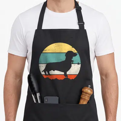 Puppy wiener apron - Grey dachshund silhouette against yellow teal white and orange retro stripes — shown on a black cotton canvas apron against a white background