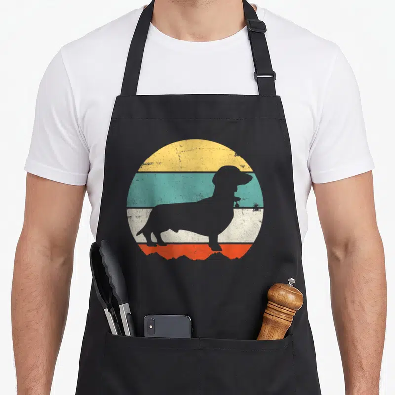 Puppy wiener apron - Grey dachshund silhouette against yellow teal white and orange retro stripes — shown on a black cotton canvas apron against a white background