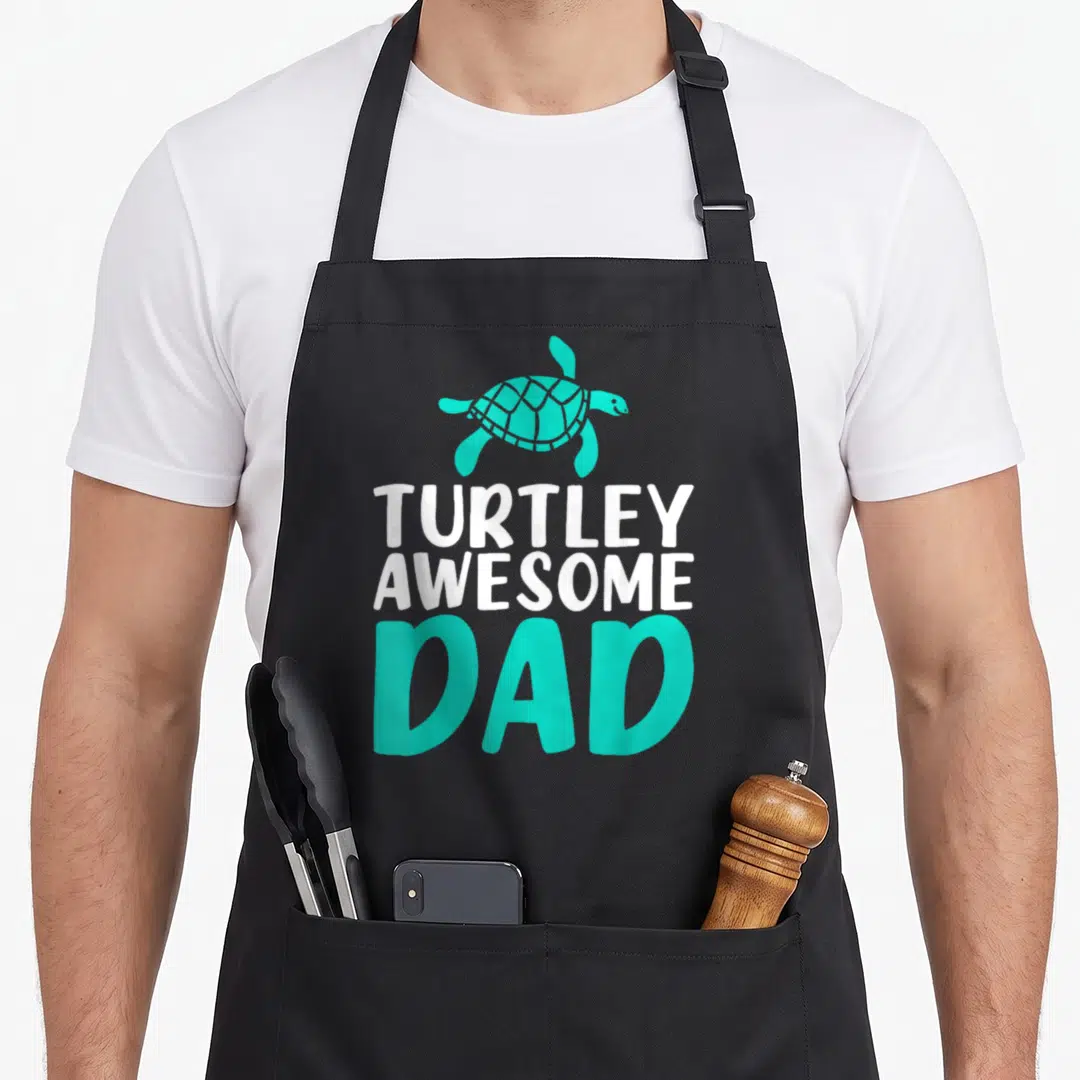 Turtley sea turtle dad apron - Teal turtle illustration above white and teal text reading TURTLEY AWESOME DAD — shown on a black cotton canvas apron against a white background