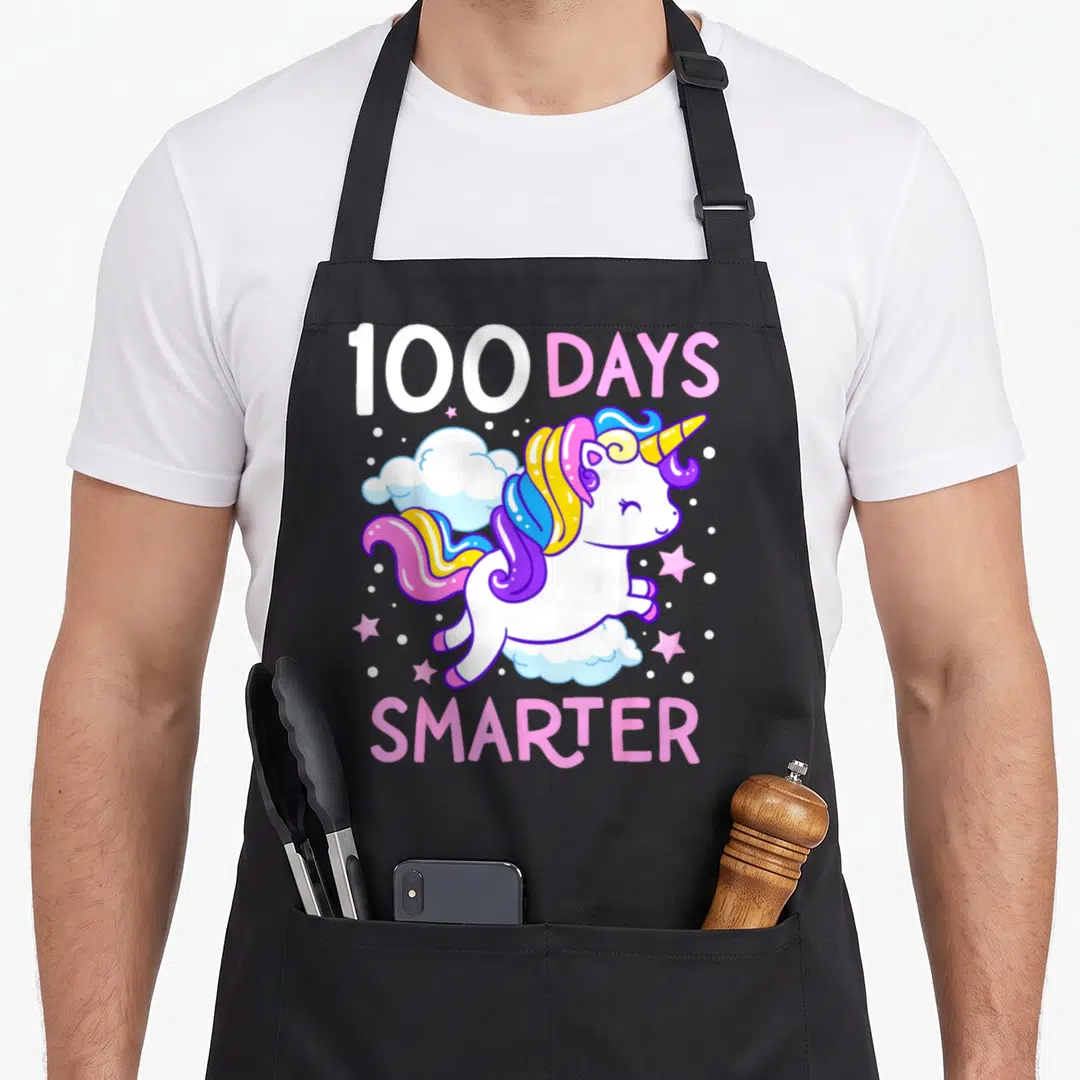 100th unicorn aprons - Cartoon unicorn with rainbow mane jumping through clouds under 100 DAYS SMARTER text — shown on a black cotton canvas apron against a white background