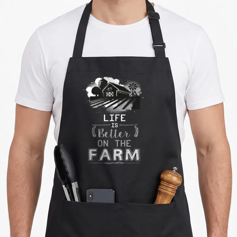Support farming apron - Black farm graphic with barn silo windmill and LIFE IS Better ON THE FARM text flanked by wheat — shown on a black cotton canvas apron against a white background