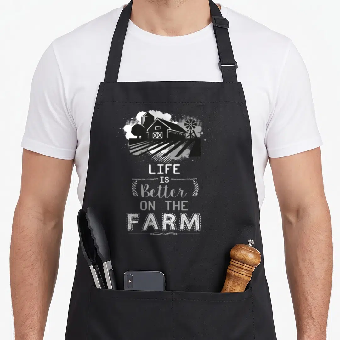 Support farming apron - Black farm graphic with barn silo windmill and LIFE IS Better ON THE FARM text flanked by wheat — shown on a black cotton canvas apron against a white background