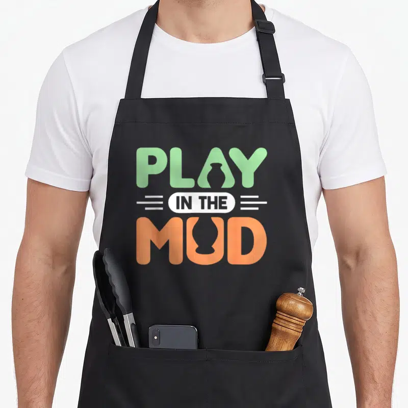Pottery ceramic clay apron - Stacked text reading PLAY IN THE MUD in green and orange with vessel silhouettes inside — shown on a black cotton canvas apron against a white background