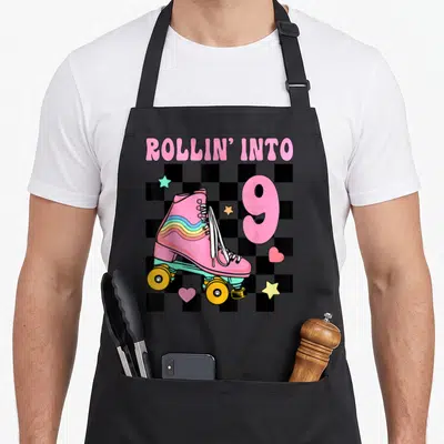 Rolling roller skating birthday apron - Pink roller skate with ROLLIN INTO 9 text on checkerboard — shown on a black cotton canvas apron against a white background
