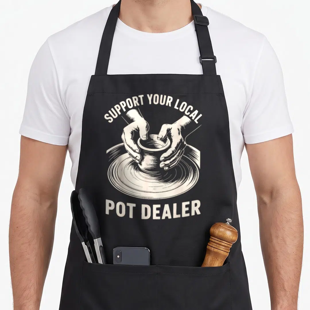 Support dealer apron - Beige line art of hands shaping clay on a wheel with arched text above and straight text below — shown on a black cotton canvas apron against a white background