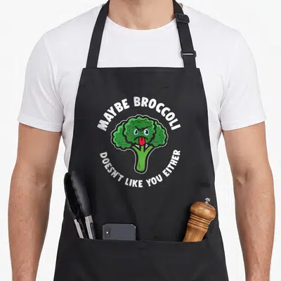 Broccoli vegetarian apron - Cartoon green broccoli sticking out a red tongue with arched white text reading maybe — shown on a black cotton canvas apron against a white background