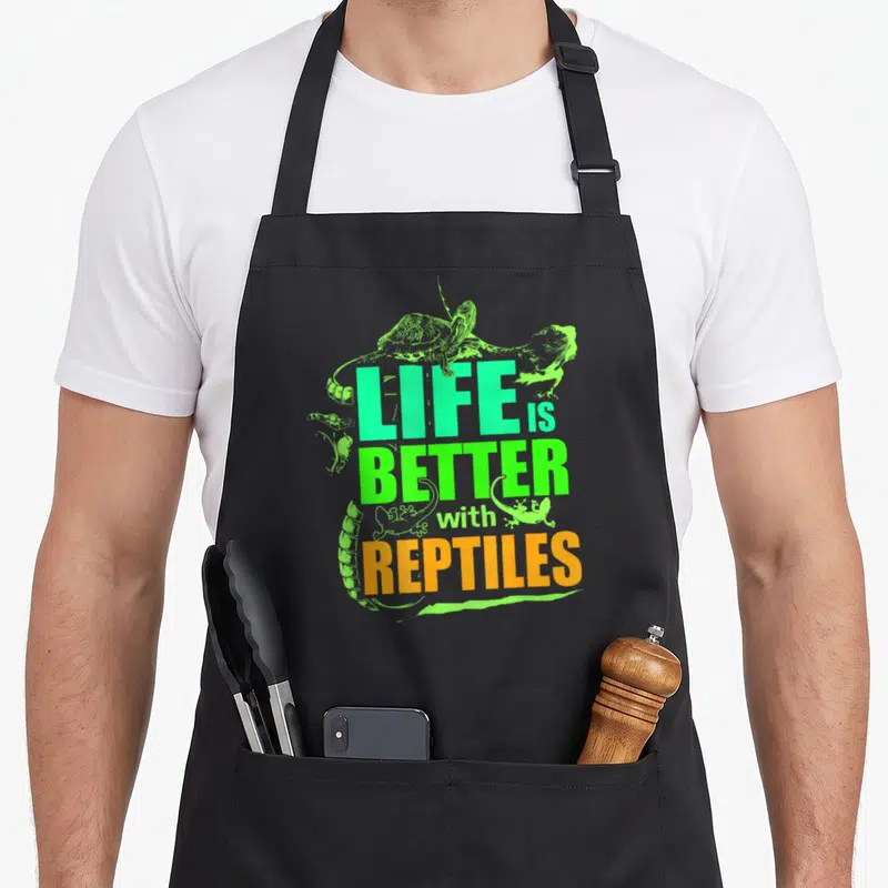 Better reptile leopard apron - Stacked LIFE BETTER REPTILES text in turquoise lime and orange with cartoon reptiles — shown on a black cotton canvas apron against a white background