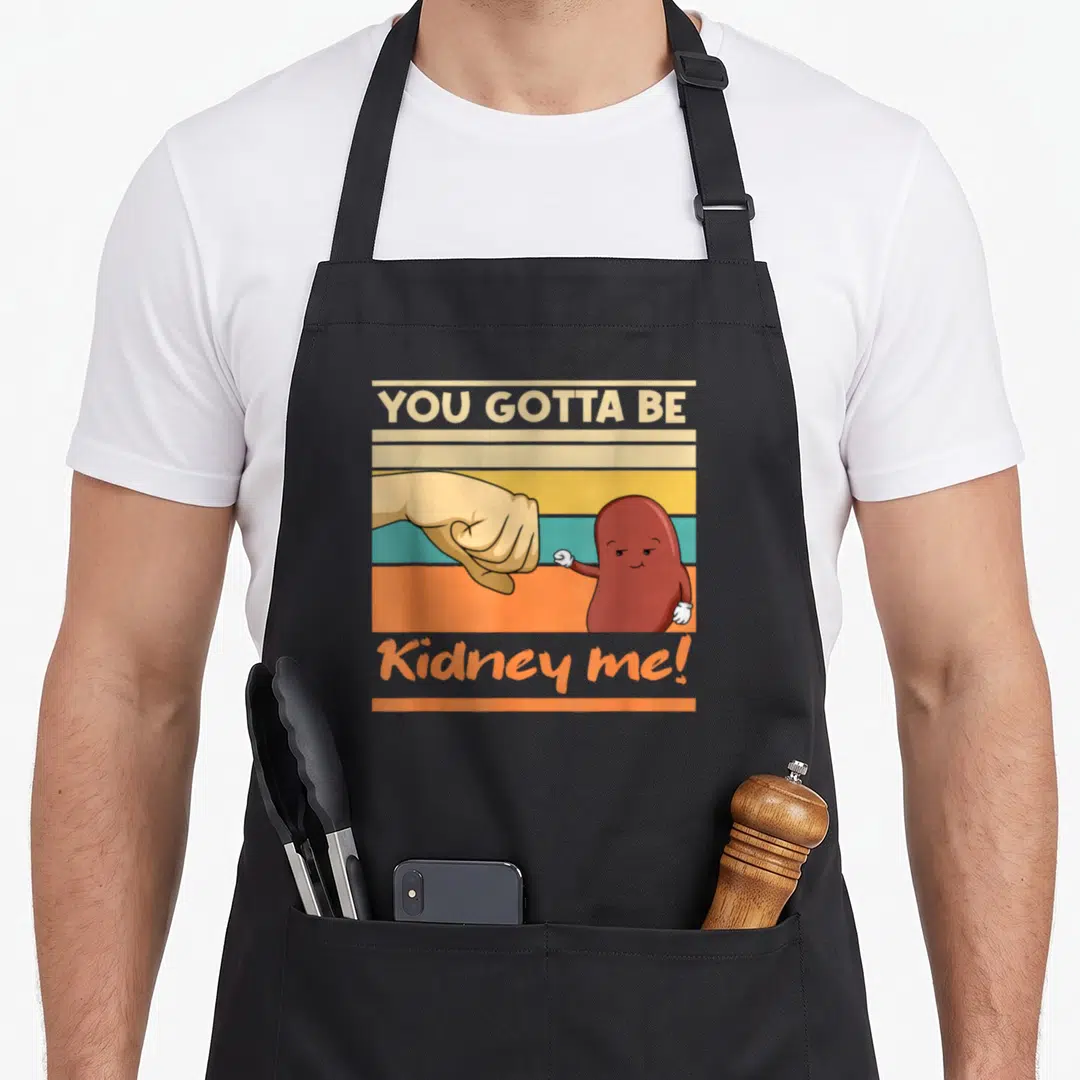 Kidney pun apron - Fist bumping cartoon kidney with YOU GOTTA BE Kidney me text on stripes — shown on a black cotton canvas apron against a white background