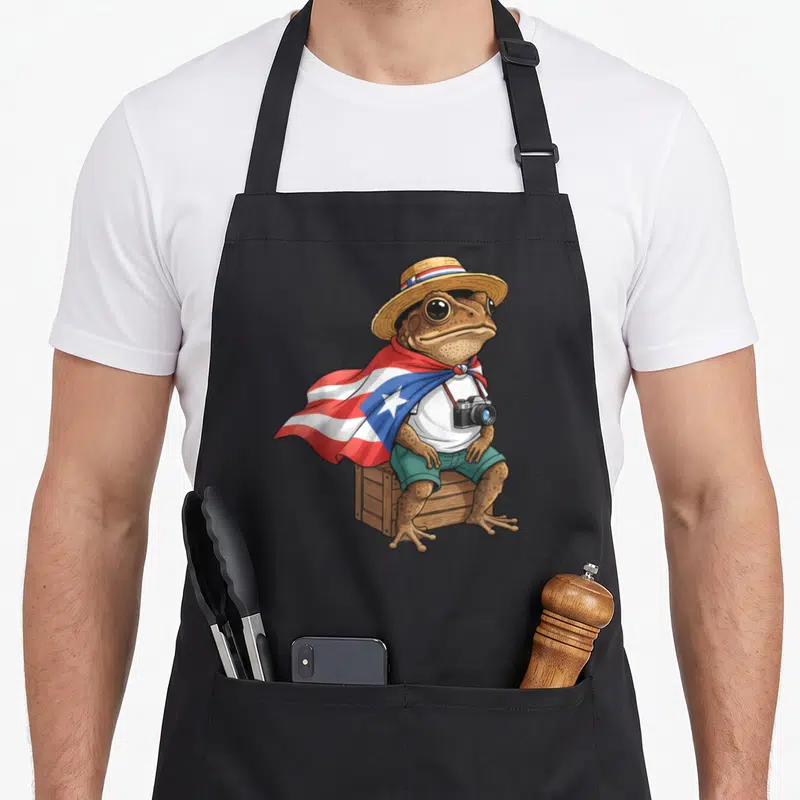 Flag frog aprons - Cartoon brown toad in flag cape holding camera sitting on crate — shown on a black cotton canvas apron against a white background