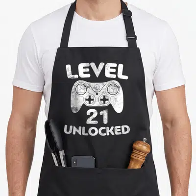 Unlocked 21st birthday apron - White distressed game controller graphic with LEVEL 21 UNLOCKED text — shown on a black cotton canvas apron against a white background