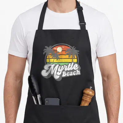 Beach 70s apron - Retro Myrtle Beach sunset graphic with palm trees and white script lettering in orange and yellow — shown on a black cotton canvas apron against a white background