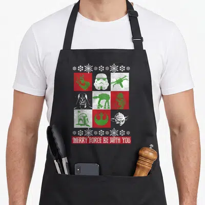 Funny force merry christmas apron - Star Wars silhouettes in red green white grid with Merry Force Be With You text — shown on a black cotton canvas apron against a white background