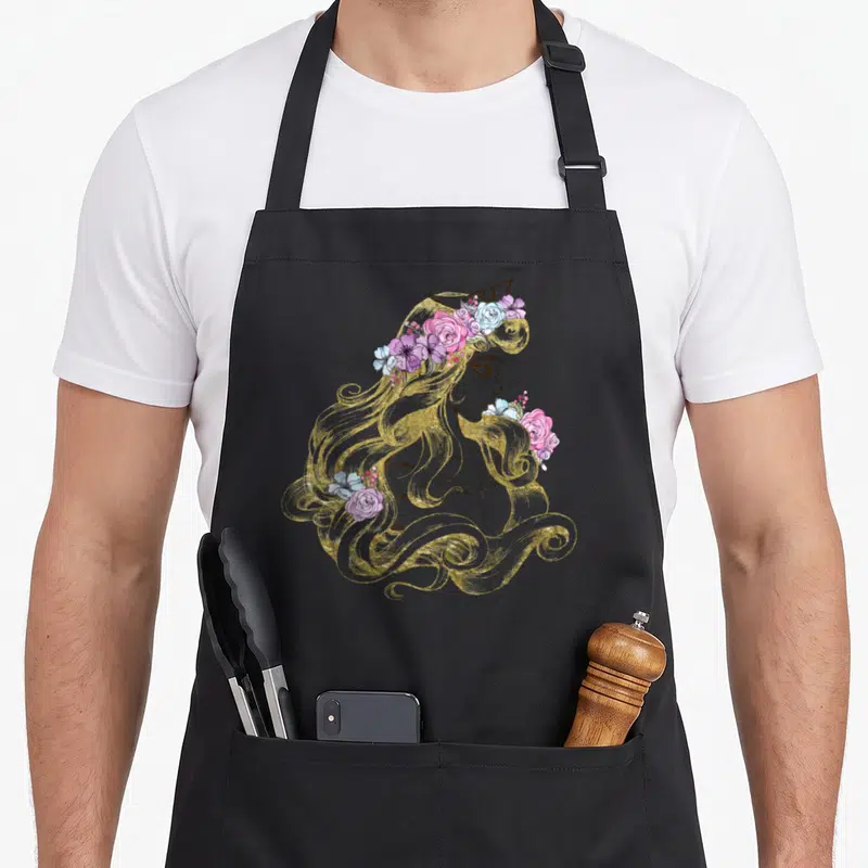 Colorful beauty apron - Line art woman profile with gold hair, crown, and pink blue purple flowers — shown on a black cotton canvas apron against a white background