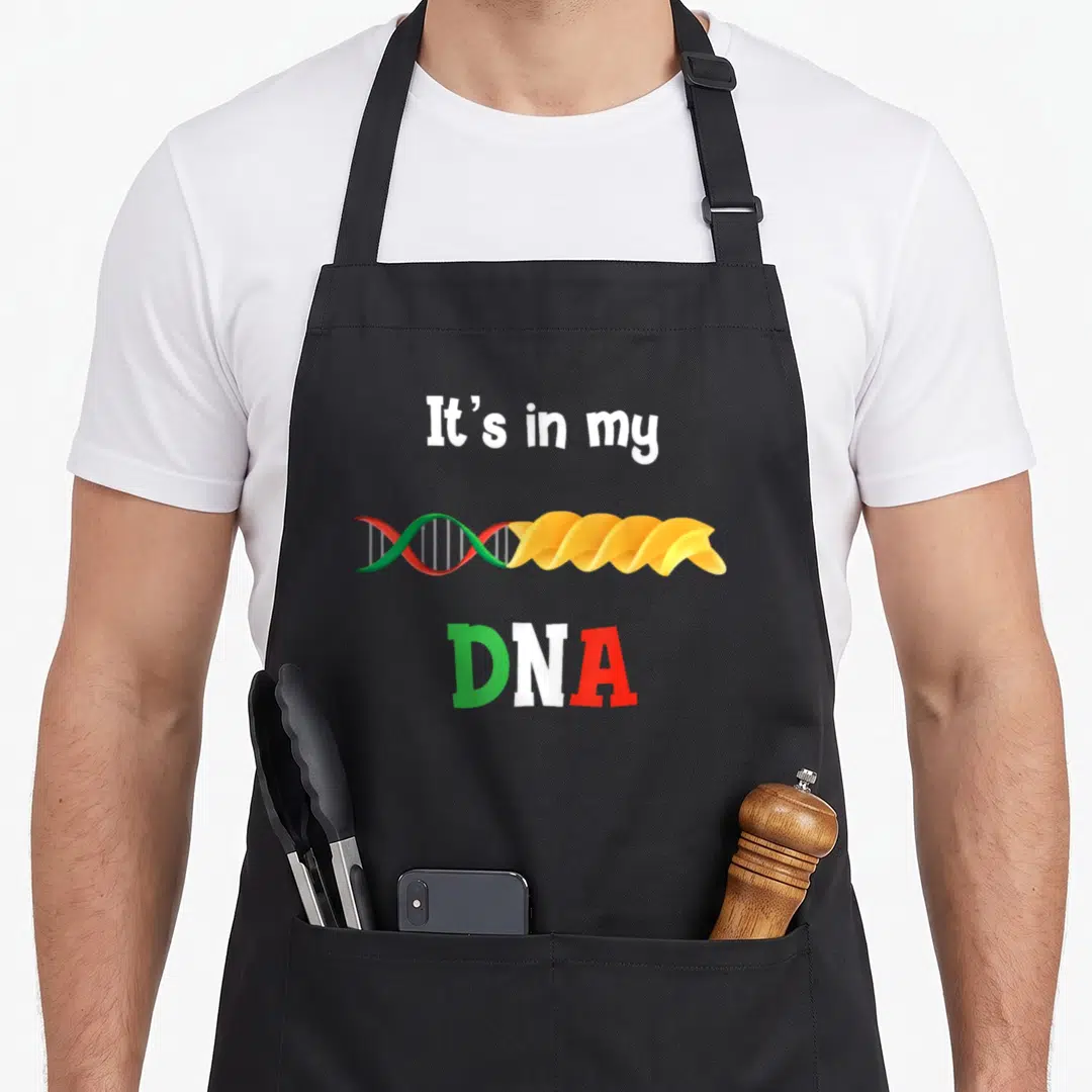 Dna pasta apron - It's in my DNA text above a helix morphing into yellow spiral pasta with green white and red letters — shown on a black cotton canvas apron against a white background