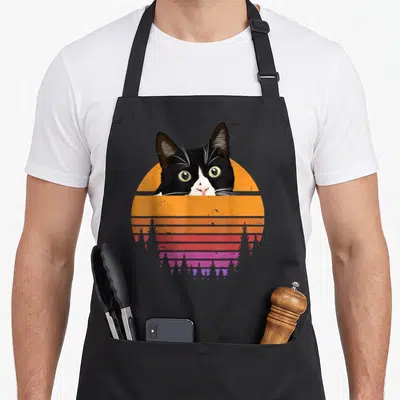 70s tuxedo apron - Cartoon tuxedo cat head over striped orange and pink sunset circle — shown on a black cotton canvas apron against a white background