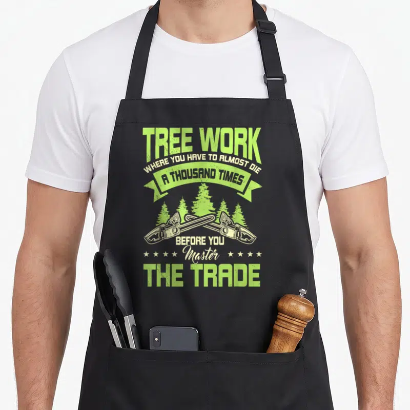 Arboriculturist surgeon apron - Crossed chainsaws and pine trees with text Tree Work Where You Have To Almost Die — shown on a black cotton canvas apron against a white background