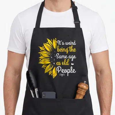 Vintage age weird aprons - Yellow sunflower graphic beside stacked white and yellow text reading It's weird being — shown on a black cotton canvas apron against a white background