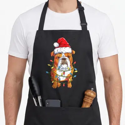 Santa english chef aprons - Cartoon bulldog in red Santa hat wrapped in red yellow blue and white Christmas lights — shown on a black cotton canvas apron against a white background