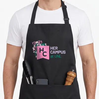 Unl campus apron - Pink banner with HC letters, flowers, and HER CAMPUS at UNL text in purple and teal — shown on a black cotton canvas apron against a white background