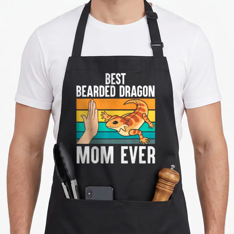 Bearded mother's day aprons - Cartoon bearded dragon high-fiving a hand on orange, yellow, and teal stripes with text — shown on a black cotton canvas apron against a white background