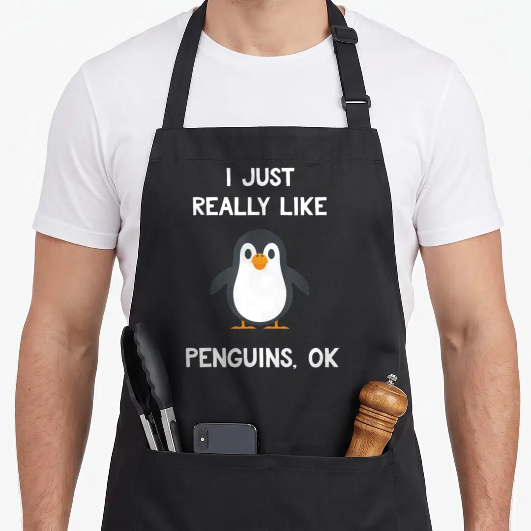 Funny penguin aprons - Cartoon penguin between text I JUST REALLY LIKE PENGUINS, OK in white grey and orange — shown on a black cotton canvas apron against a white background