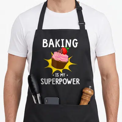 Superpower baker cupcake apron - Cartoon cupcake with pink frosting and strawberry inside yellow burst with white text — shown on a black cotton canvas apron against a white background