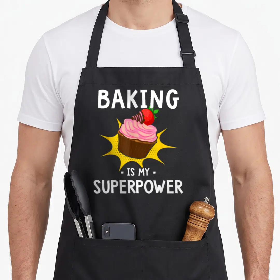 Superpower baker cupcake apron - Cartoon cupcake with pink frosting and strawberry inside yellow burst with white text — shown on a black cotton canvas apron against a white background