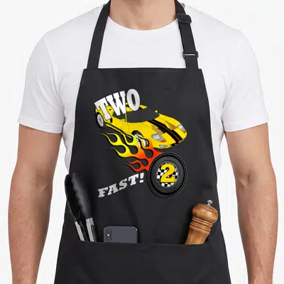 Race racing car apron - Yellow cartoon car with flames and text TWO FAST! and number 2 in tire — shown on a black cotton canvas apron against a white background