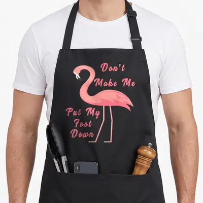 Foot flamingo apron for grilling - Pink cartoon flamingo with text Don't Make Me Put My Foot Down beside its neck — shown on a black cotton canvas apron against a white background