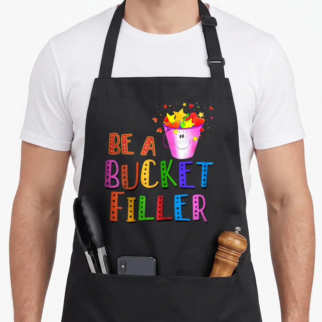 Growth bucket apron - Smiling pink bucket with yellow stars and red hearts above stacked colorful text BE A BUCKET — shown on a black cotton canvas apron against a white background