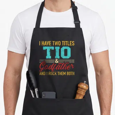 Title godfather apron - Stacked yellow teal and red text reading I have two titles Tio and Godfather — shown on a black cotton canvas apron against a white background