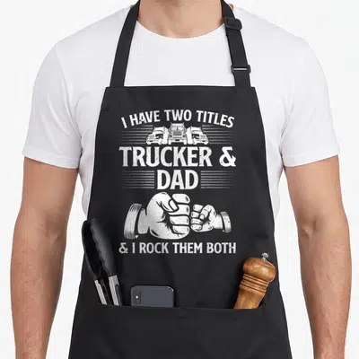 Driving truck apron - White distressed text I Have Two Titles Trucker & Dad with three trucks and a fist bump graphic — shown on a black cotton canvas apron against a white background