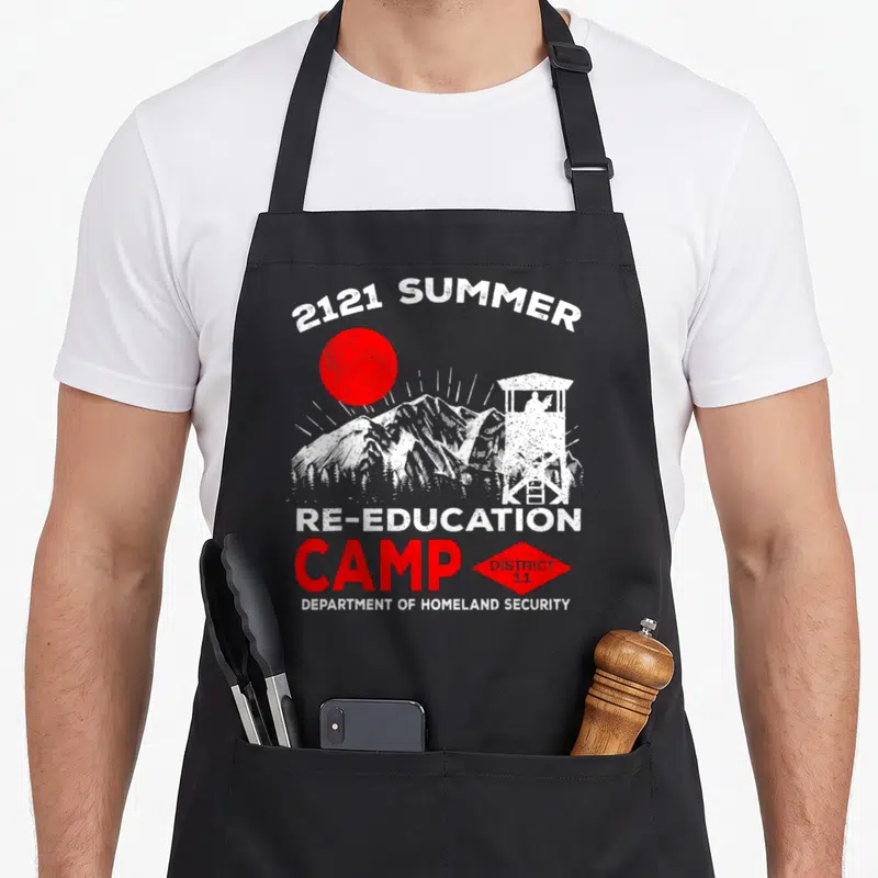 Reeducation camp chef apron - Distressed graphic of red sun, white mountains, watchtower, and text 2121 Summer — shown on a black cotton canvas apron against a white background