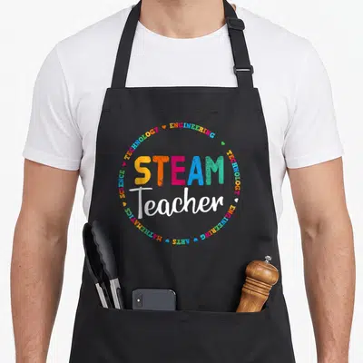 Stem teacher apron - Colorful STEAM Teacher text surrounded by Engineering Technology Science Math Arts words and hearts — shown on a black cotton canvas apron against a white background