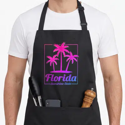 Sunshine state florida apron - Retro graphic of three pink and blue palm trees on an island with Florida text — shown on a black cotton canvas apron against a white background