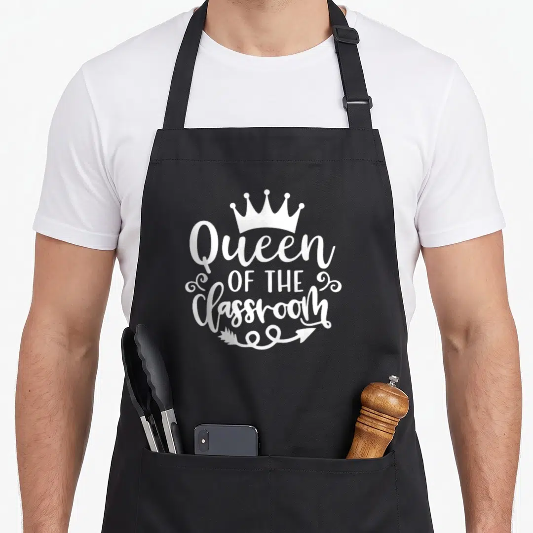 Education classroom aprons - White crown, stacked Queen OF THE Classroom text, swirls, and arrow graphic — shown on a black cotton canvas apron against a white background