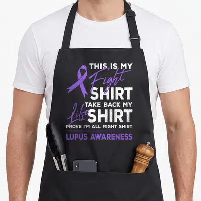 Fight lupus ribbon apron - Purple awareness ribbon next to stacked white and purple text reading This Is My Fight Shirt — shown on a black cotton canvas apron against a white background