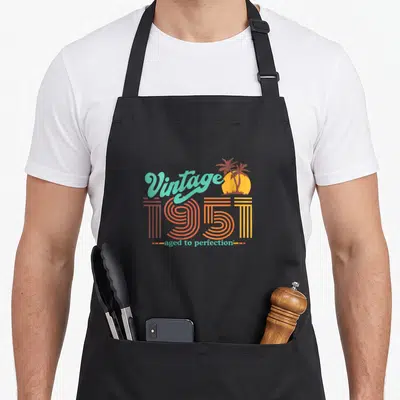 Funny aged 70th birthday apron - Retro 1951 text with palm trees and sun in teal orange yellow brown — shown on a black cotton canvas apron against a white background