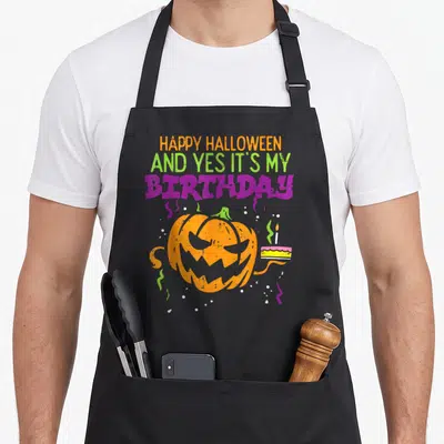 Vintage birthday funny halloween apron - Orange pumpkin holding cake under HAPPY HALLOWEEN AND YES IT'S MY BIRTHDAY text — shown on a black cotton canvas apron against a white background